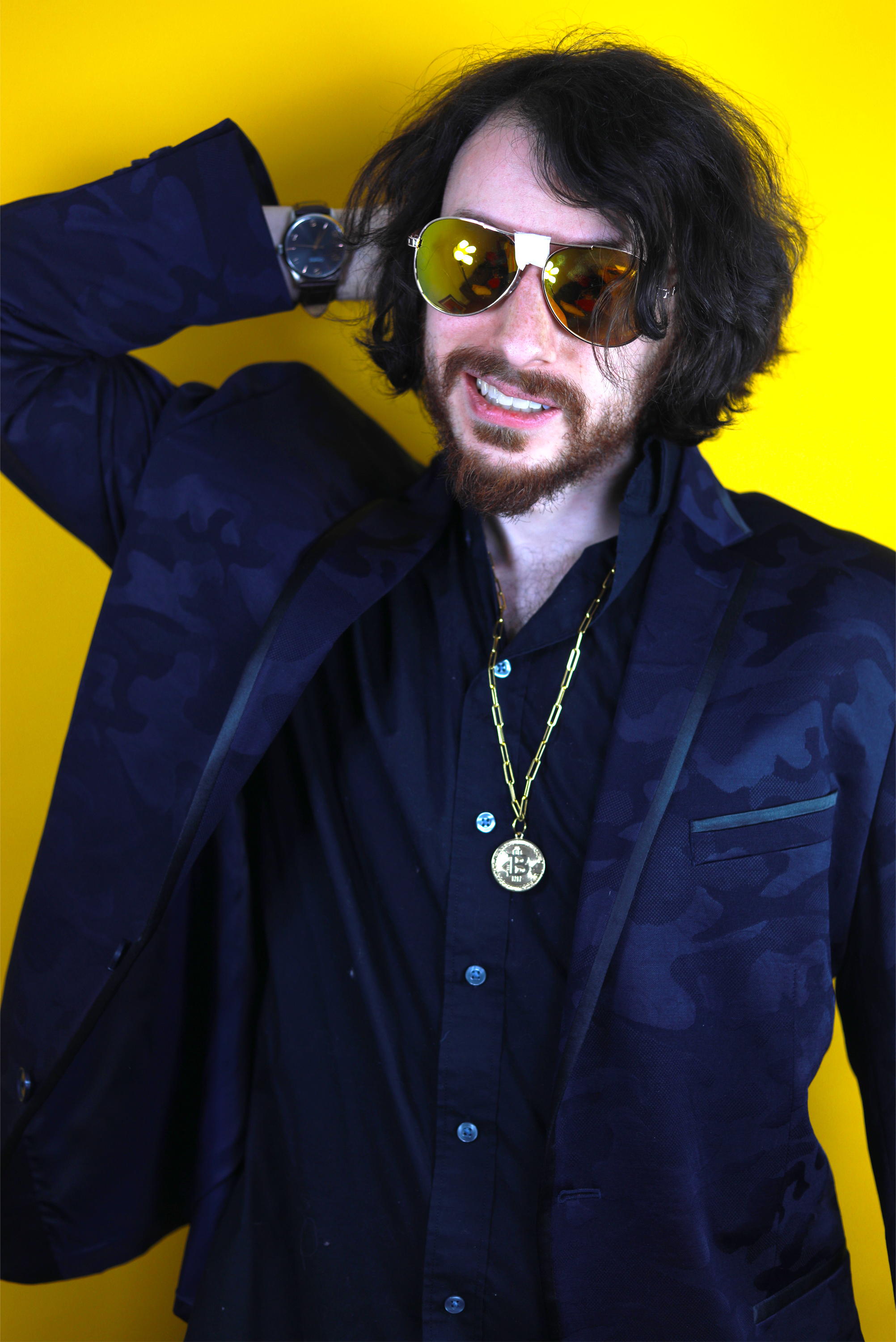 Rito wearing a blue suit with a bitcoin necklace with his arm behind with head against a yellow background