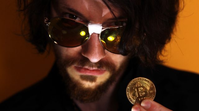 Rito holding a Bitcoin coin in a shadowy background wearing sunglasses with tape in the middle